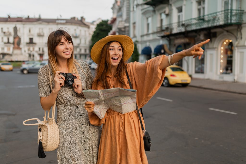 stylish young women traveling together in Europe dressed in spring trendy dresses and accessories smiling happy friends having fun taking photo on camera holding map
